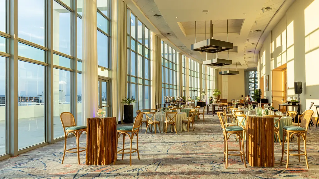The interior of a spacious, high-ceilinged event hall bathed in warm afternoon sunlight. A massive, curved wall of floor-to-ceiling windows on the left looks out toward the coast. The room is set up for a reception with patterned carpeting, round dining tables, and rustic wood high-top tables paired with woven barstools.