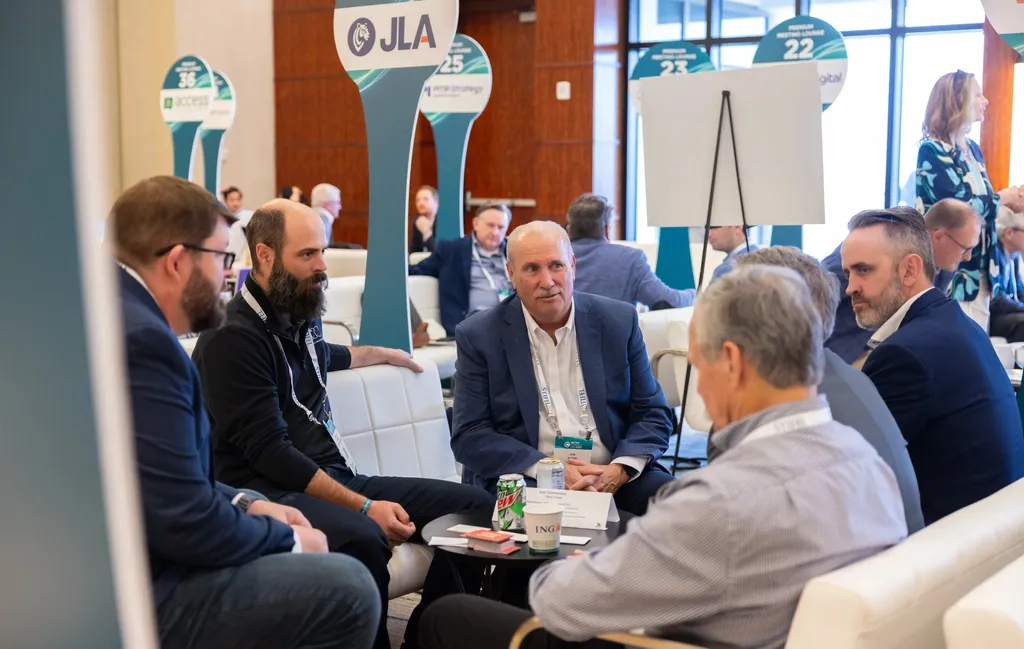 A close-up shot of a group of professionals engaged in a focused conversation at a networking event. They are seated in white leather armchairs around a small black coffee table holding drinks and business cards. In the background, tall teal signage with corporate logos like 