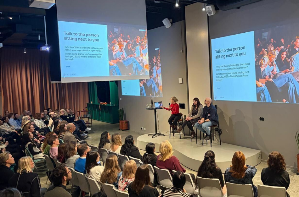 A panel discussion is taking place in a well-lit room, with audience members seated and focused on speakers at the front. There is a presentation displayed on a screen behind the speakers, emphasizing engagement among attendees.