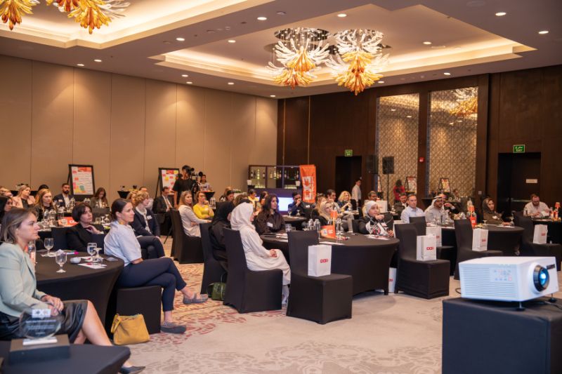 A conference room filled with attendees seated at tables, some taking notes or looking towards a projector screen. The decor includes ornate light fixtures.