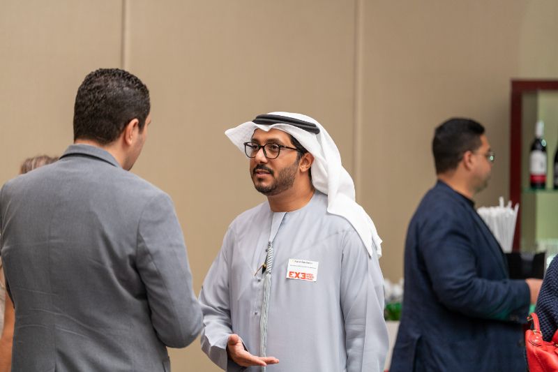 Two men are engaged in a conversation at a social event. One man is wearing traditional Emirati attire, including a white kandura and ghutrah, while the other is dressed in business attire. The background features other attendees and some refreshments.