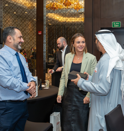 Three individuals engaged in conversation at a social event, with one man wearing a suit and tie, a woman in a blazer and leather skirt, and another man in traditional Emirati attire.