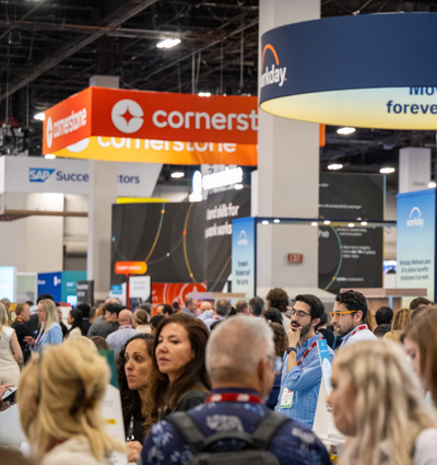 A crowded exhibition space with numerous attendees interacting, featuring banners and logos from various companies like Cornerstone and Workday.