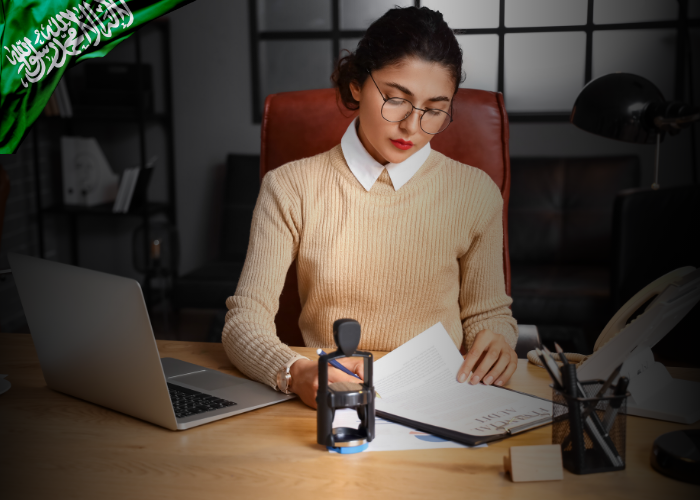 Alt Text: A professional woman wearing glasses and a beige sweater sits at a desk in a modern office, reviewing documents beside an open laptop. A stamp device and office supplies are placed on the desk. In the top left corner, a green Saudi Arabian flag graphic is overlaid, suggesting official or national affiliation.