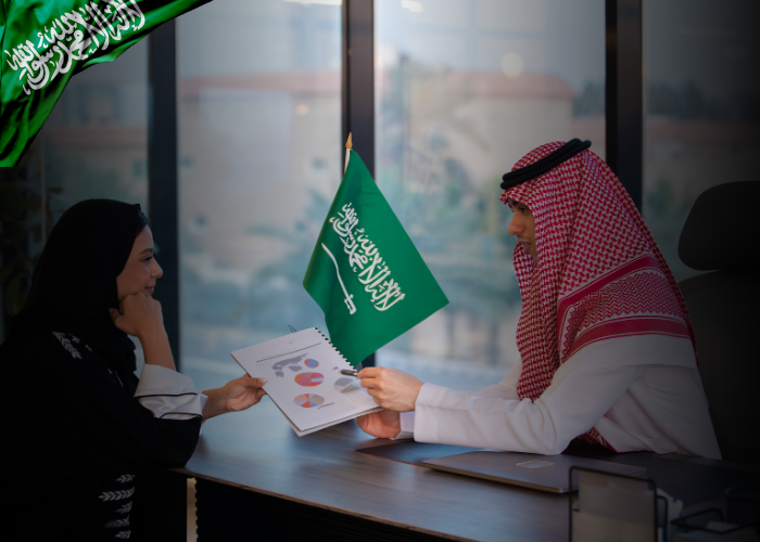 A Saudi man and woman in traditional attire are seated at a desk in a modern office, reviewing printed charts with graphs. A small Saudi Arabian flag is placed between them, symbolizing national context and collaboration. Natural light streams through large windows in the background.