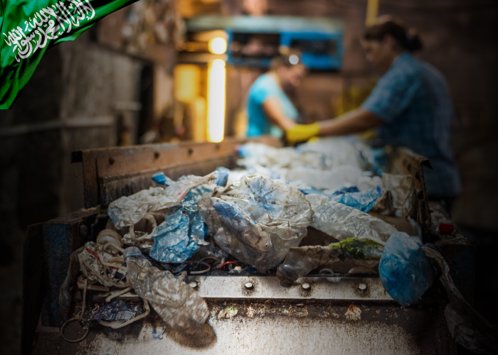 A conveyor belt filled with dirty plastic waste and debris moves through a recycling facility. In the blurred background, two workers in blue clothing sort materials by hand while wearing gloves. A Saudi Arabian flag graphic is overlaid in the top left corner, indicating the regional context of the operation. 