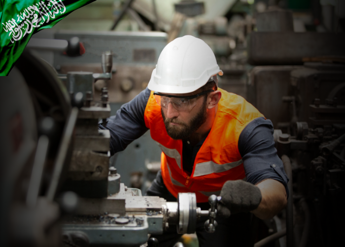 A male industrial worker wearing a white hard hat, safety goggles, and an orange reflective vest operates heavy machinery in a factory setting. He appears focused while adjusting equipment with a gloved hand. A green Saudi Arabian flag graphic is overlaid in the top left corner, indicating a national or regional industrial context.