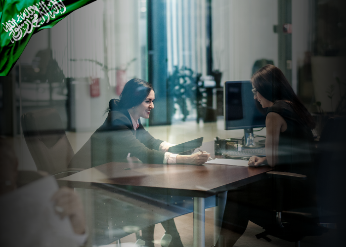 Two professional women are seated at a desk in a modern office, engaged in a discussion with documents and a tablet in hand. The atmosphere is bright and collaborative, with glass walls and contemporary furnishings. A Saudi Arabian flag graphic is overlaid in the top left corner, suggesting a national or regional business context.