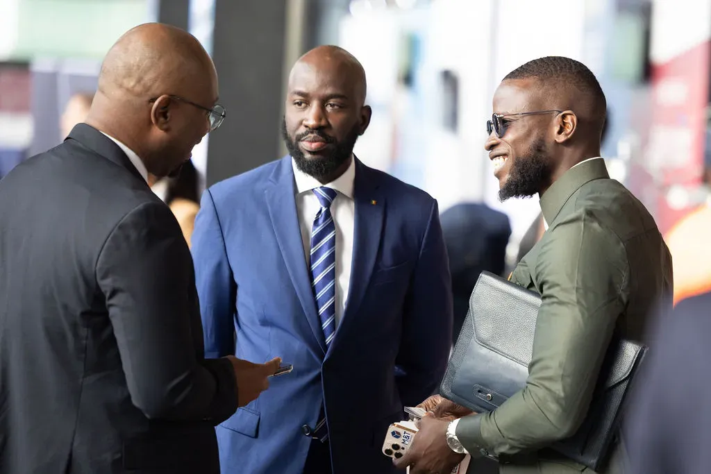A three-man interaction at an event, with the central figure in a dark blue suit and patterned tie engaged in conversation with a colleague in a olive green outfit holding a leather portfolio on the right.