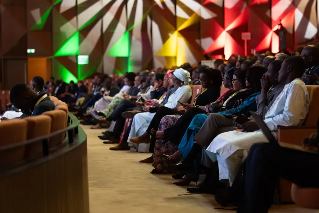 An audience seated in rows at a conference auditorium, illuminated by red and green stage lights during an event.