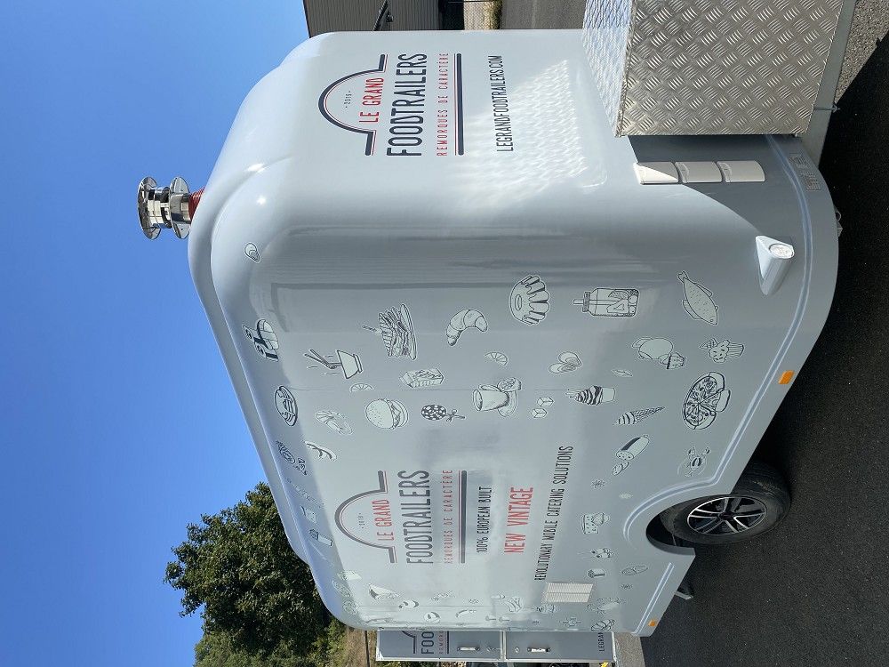 Side panel of Le Grand Foodtrailers NEW Vintage trailer featuring brand logo, slogan ‘100% European Built’, and food-themed illustrations under a clear blue sky.