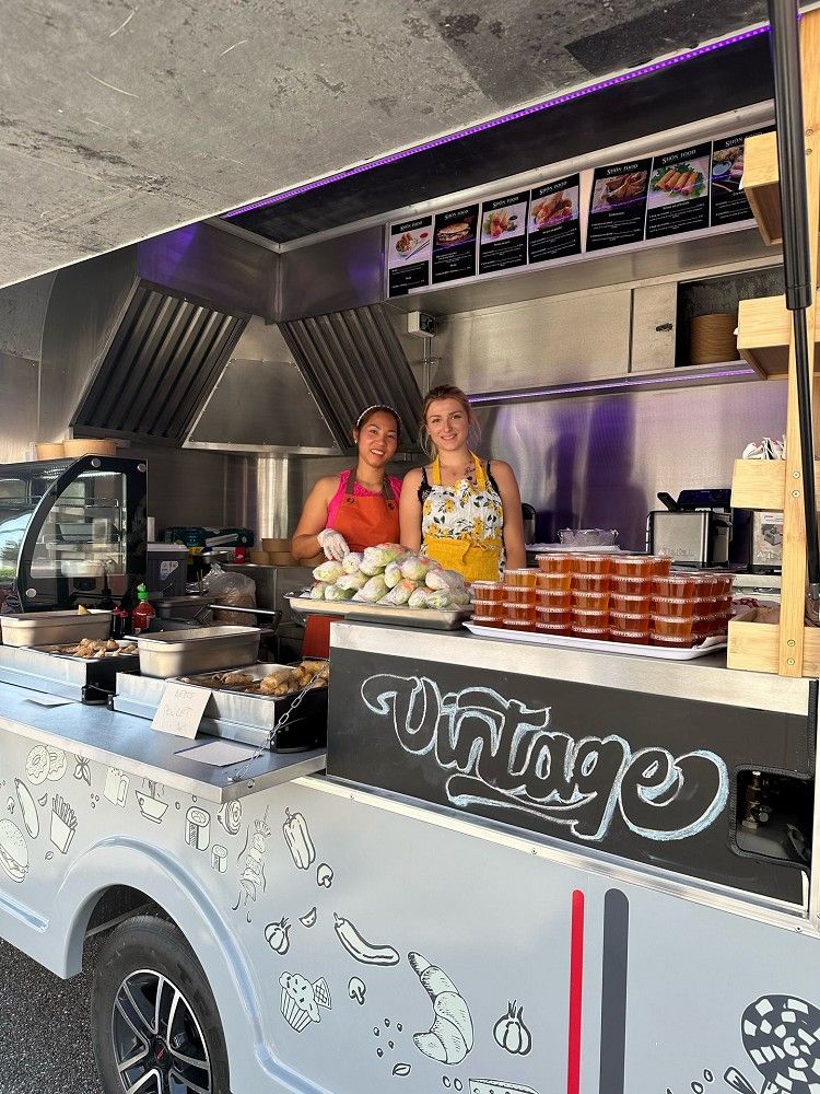 Inside the Le Grand NEW Vintage food trailer, two smiling women serve freshly made meals from a stainless-steel kitchen setup with spring rolls and condiments on display.