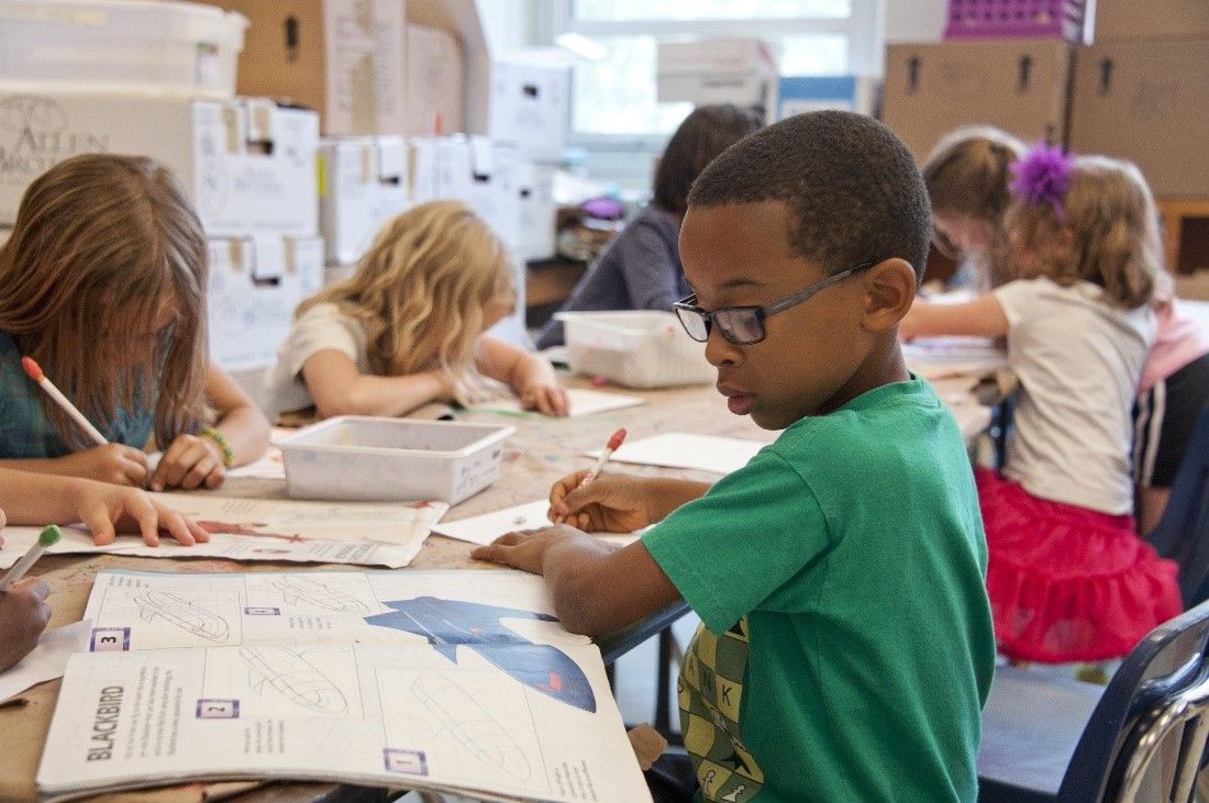 Picture of young boy with glasses drawing and looking at a magazine. 