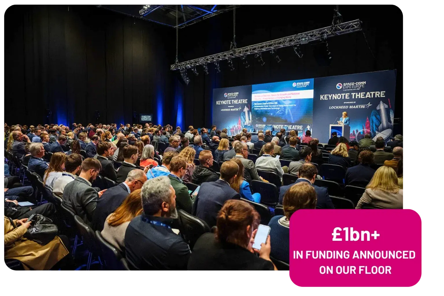 A large audience seated in a keynote theatre at a conference, with a presenter on stage and various screens displaying information.