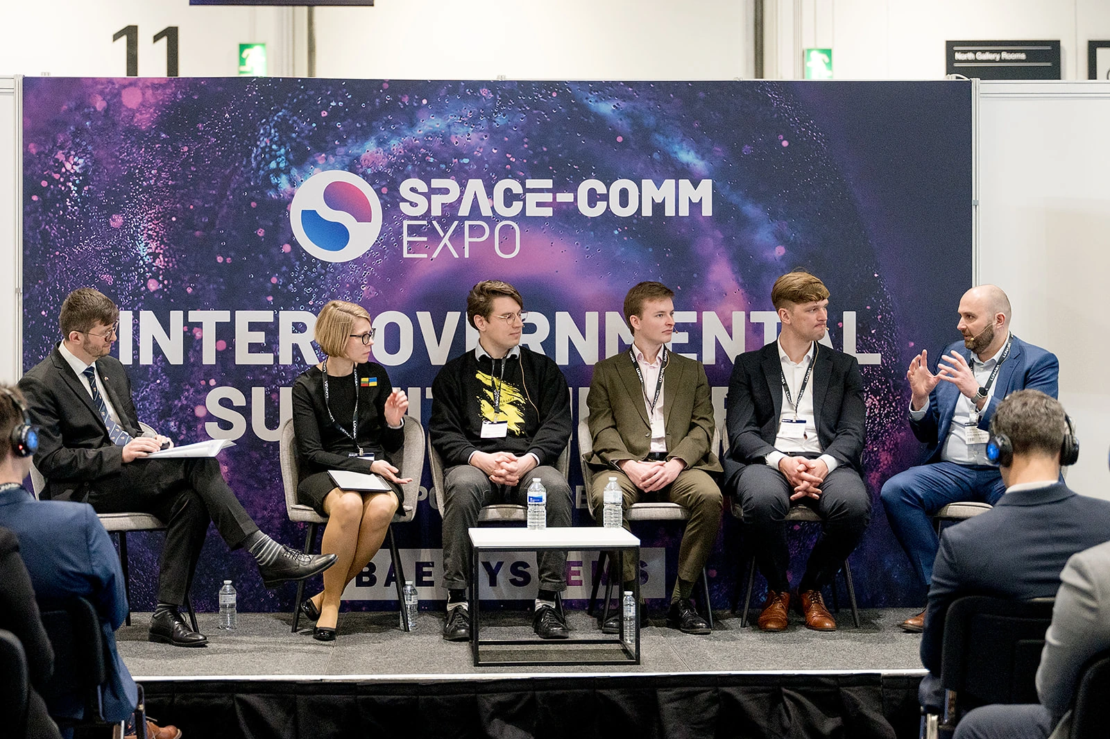 A panel discussion at the Space-Comm Expo featuring five speakers sitting on stage, with an audience in the foreground.