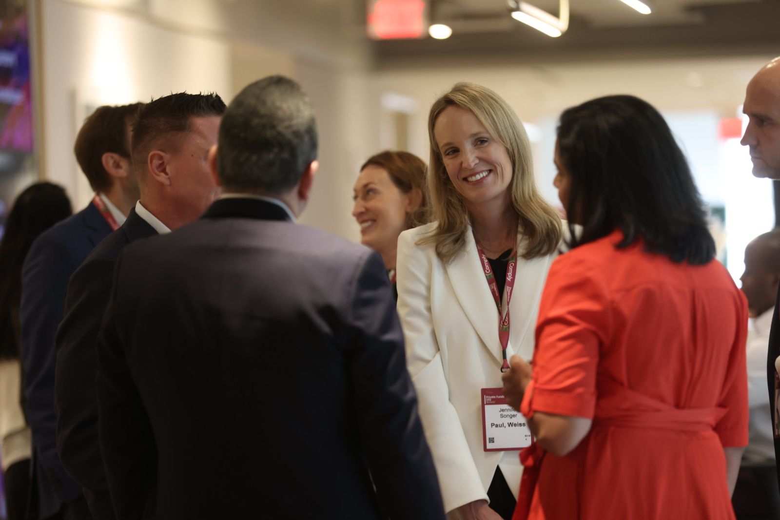 Professionals wearing conference badges converse at an indoor networking event, led by a smiling woman in white.