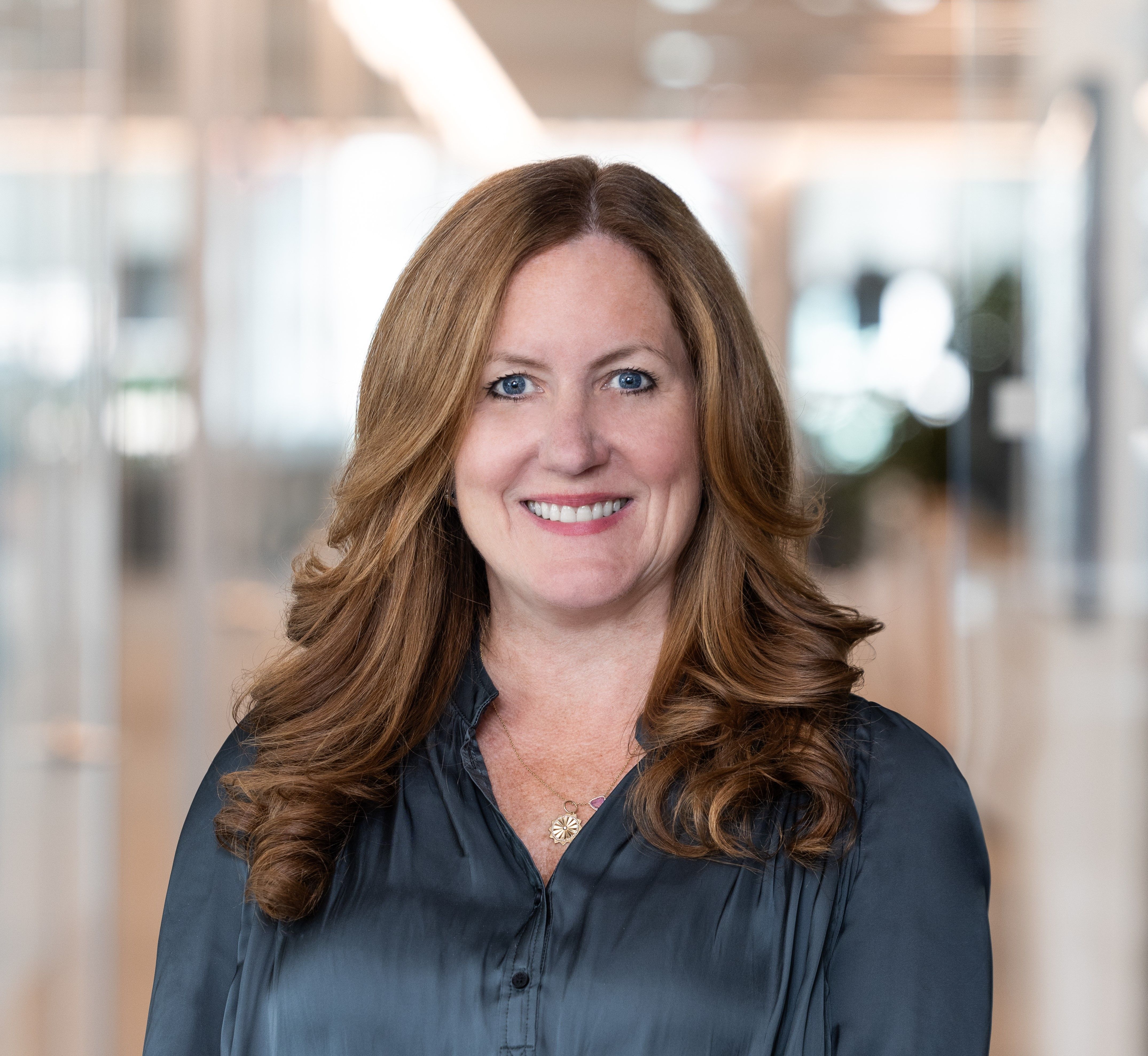 Professional headshot of a smiling woman with long auburn hair, wearing a dark blouse in a bright office.