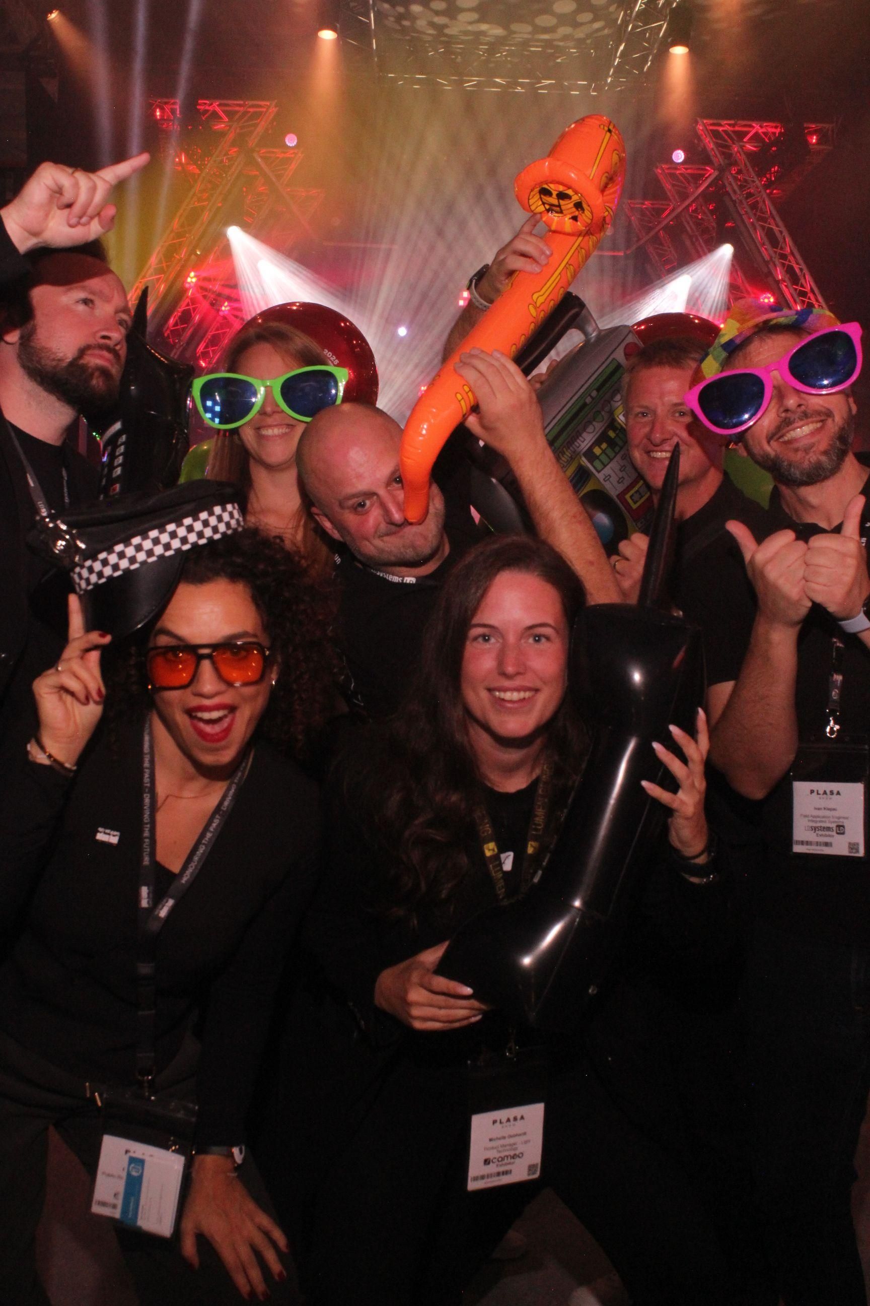 Group of PLASA Show London 2025 attendees pose for a fun photo at the after-hours party, wearing colourful accessories and holding inflatable props under stage lighting