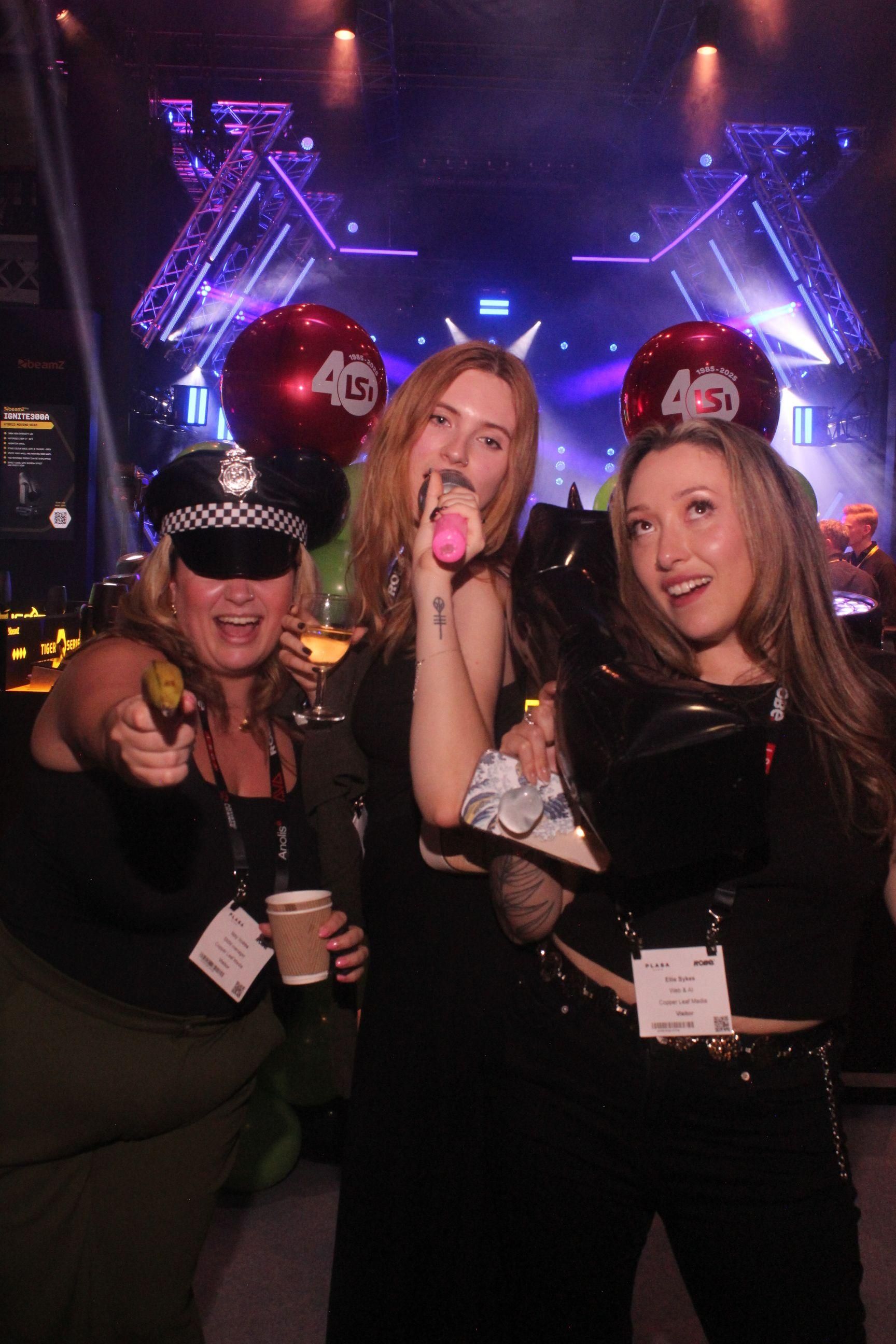 Three female visitors enjoy the PLASA Show London 2025 after-hours party, posing with props and drinks in front of the LSi 40th anniversary balloons and stage lighting display.