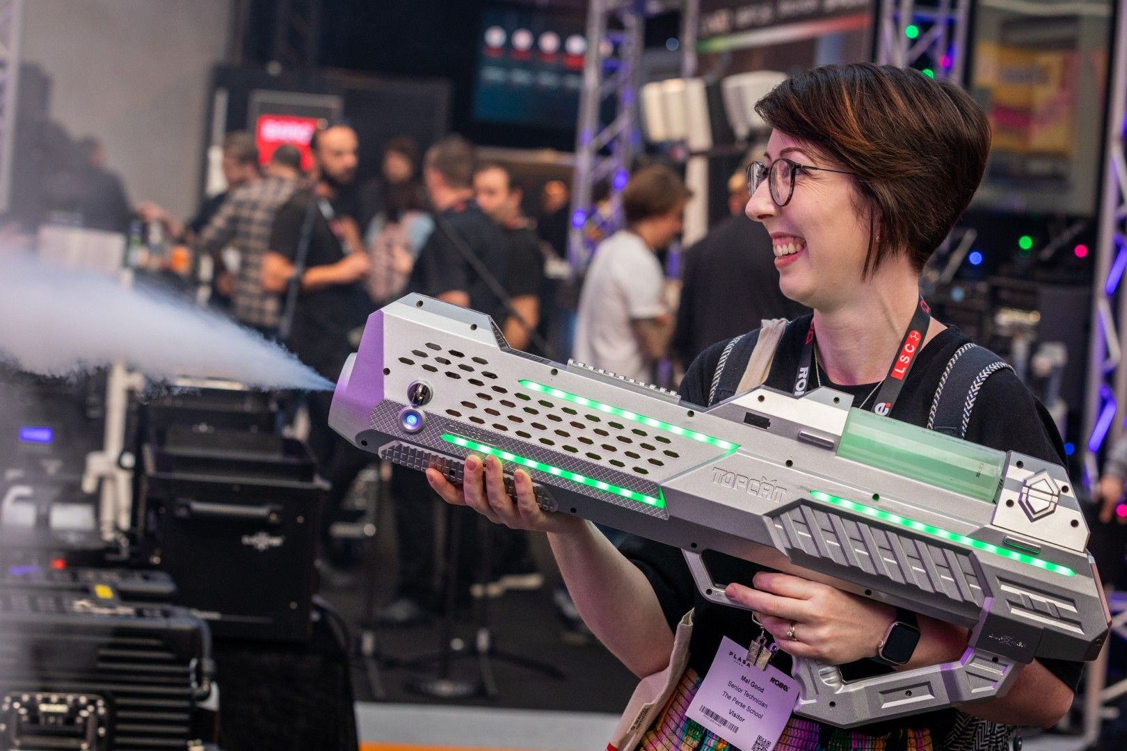 Smiling woman with short brown hair demonstrating a futuristisc hand-held fog cannon at PLASA Show London 2025.