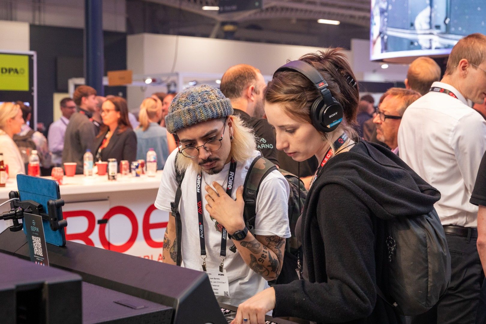 Male visitor wearing a beanie and female visitor with headphones testing audio equipment at the ROE Visual stand during PLASA Show London 2025.