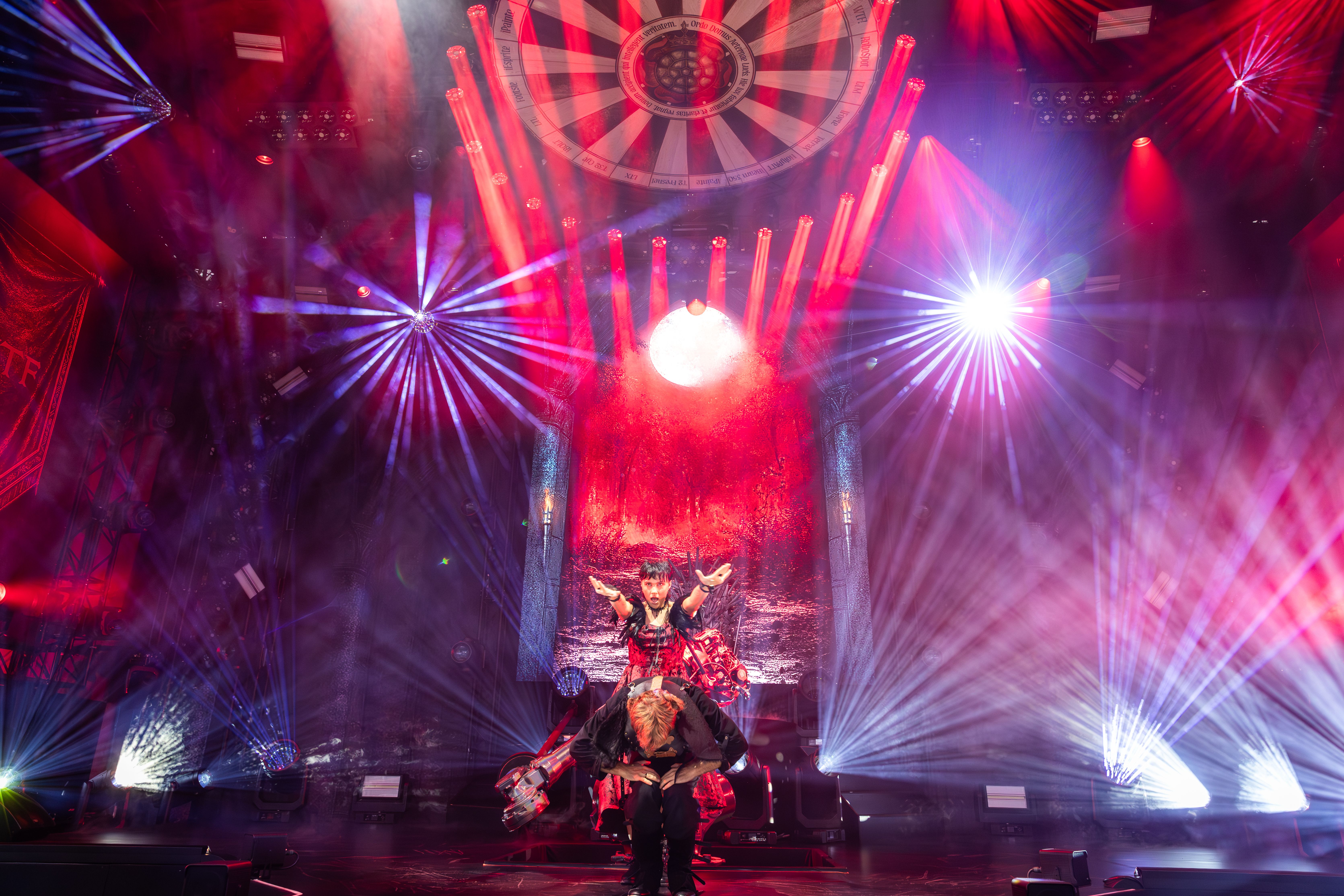 Dynamic live performance under red and white stage lighting at PLASA Show London 2025, featuring two performers in costume framed by beams of light and a medieval-inspired circular ceiling design.