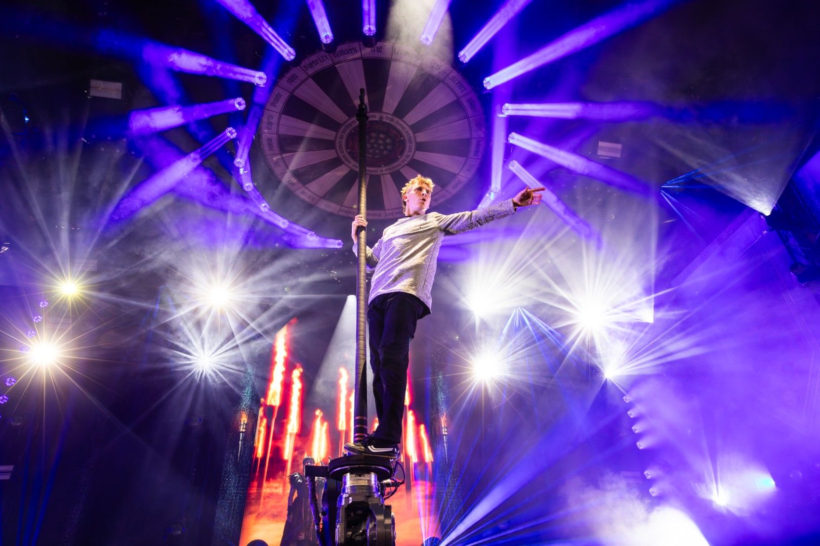Performer stands on a vertical pole surrounded by bright stage lighting effects during a live show at PLASA Show London 2025, with beams of purple and white light radiating across the stage.