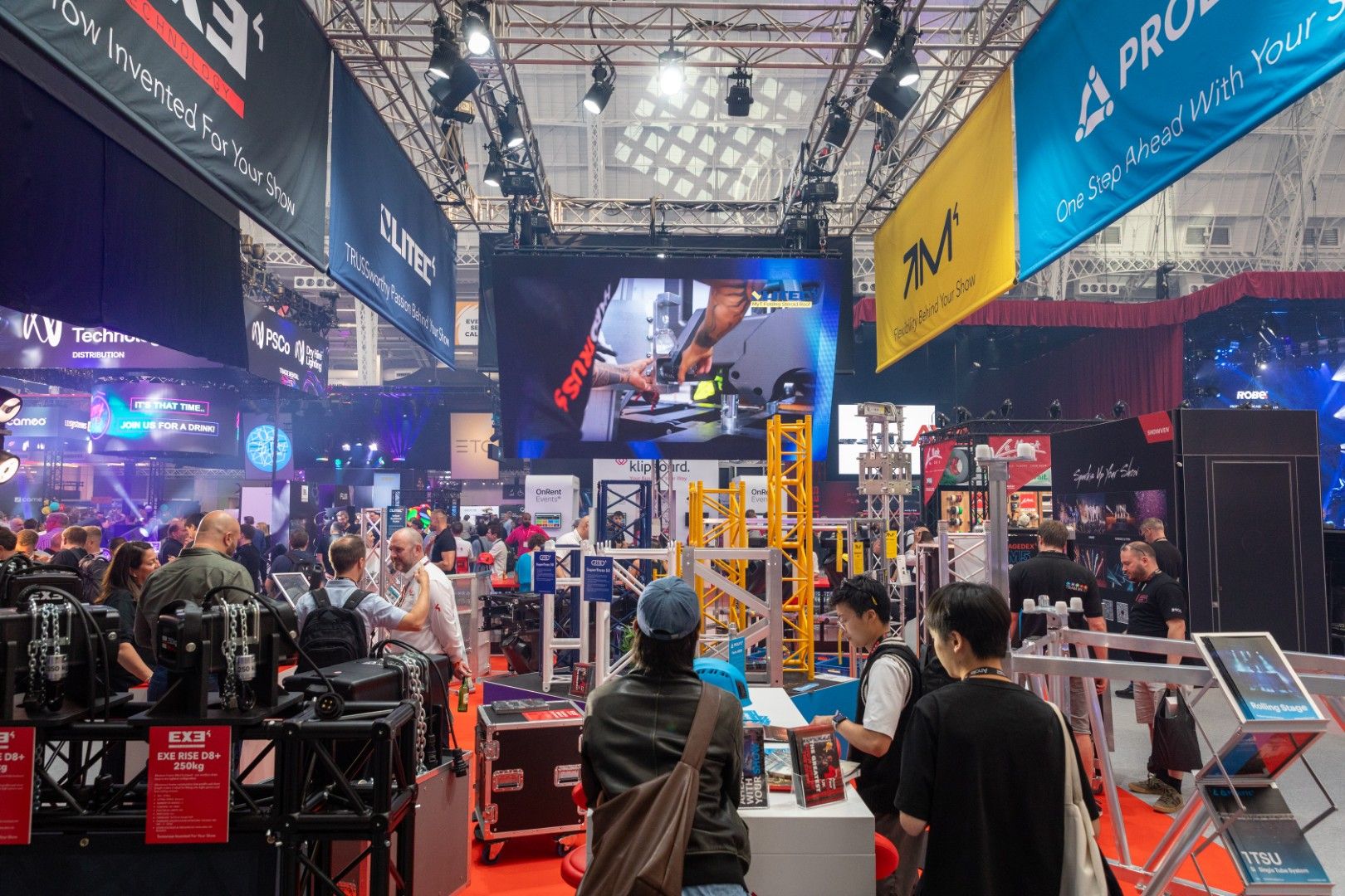 Busy exhibition floor at PLASA Show London 2025, with visitors exploring the Area Four Industries stand displaying truss structures, rigging equipment, and large video screens under banners for brands including LITEC, PROLYTE, and MILOS