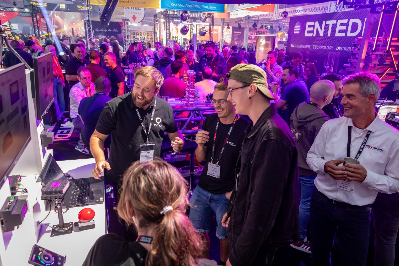 Male exhibitor in a black Entedi shirt demonstrating lighting control technology to a group of smiling visitors at PLASA Show London 2025, with the busy Entedi stand and colourful stage lighting in the background