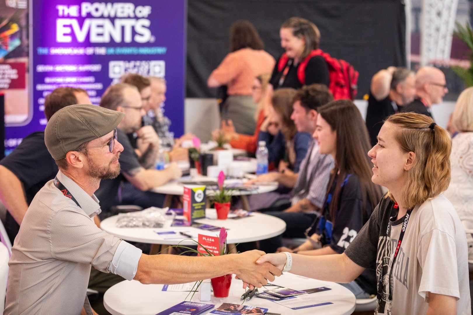A man wearing glasses and a flat cap shakes hands with a young woman during a networking session at the Careers and Networking Hub, part of PLASA Show London 2025