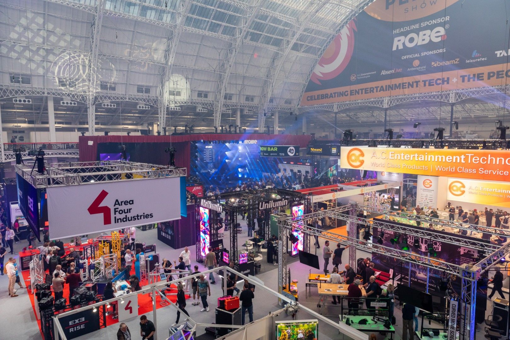 Wide view of the PLASA Show London 2025 exhibition floor at Olympia, featuring stands from A.C. Entertainment Technologies, Area Four Industries, Unilumin and Robe Lighting beneath the venue’s iconic glass roof.