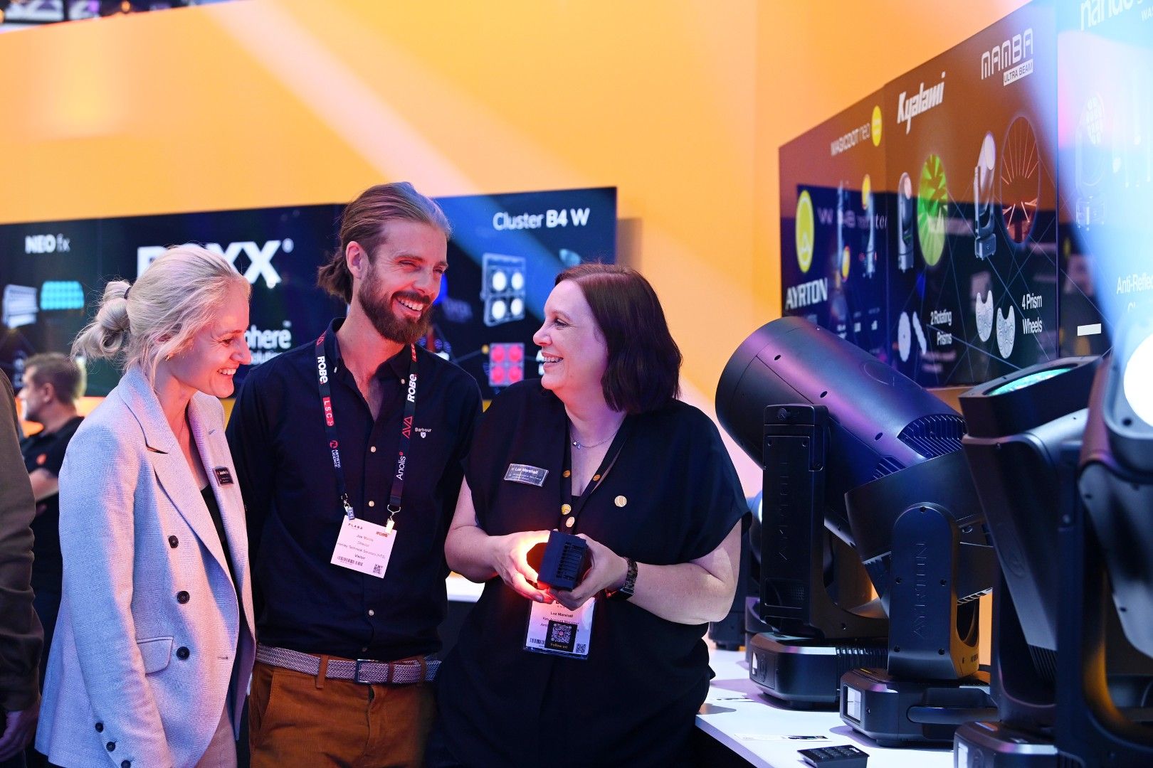 Two female visitors and a male exhibitor smiling while viewing Ayrton and ROXX lighting fixtures at PLASA Show London 2025, under vibrant display lighting on the exhibition stand.