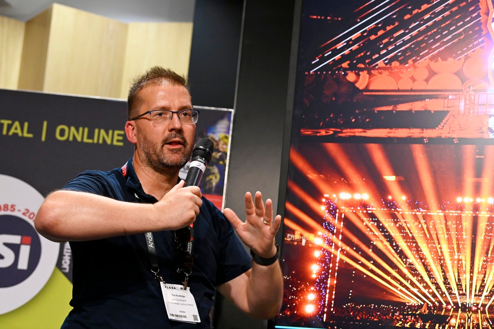 Lighting designer Tim Routledge speaking during a seminar at PLASA Show London 2025, discussing large-scale stage productions with visuals of concert lighting designs displayed behind him.