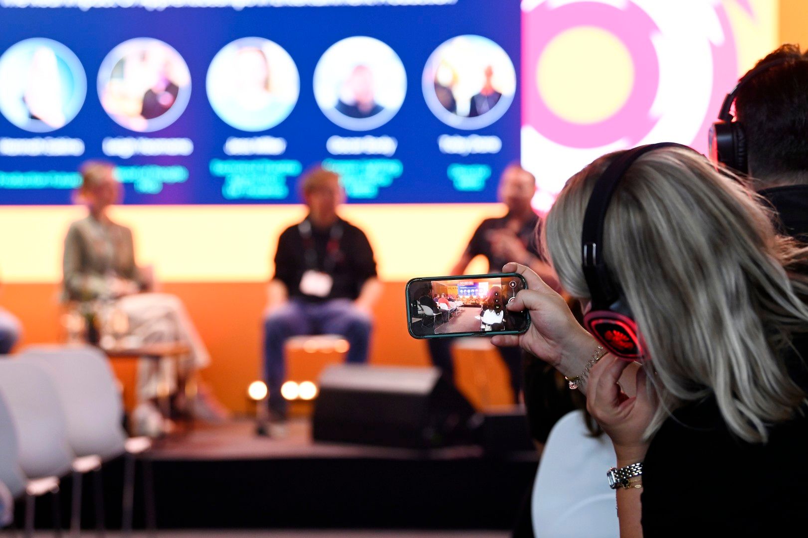 Blonde female audience member wearing headphones films a live panel discussion on their phone at PLASA Show London 2025, with speakers seated on stage in front of a bright screen displaying the session title and participant names.