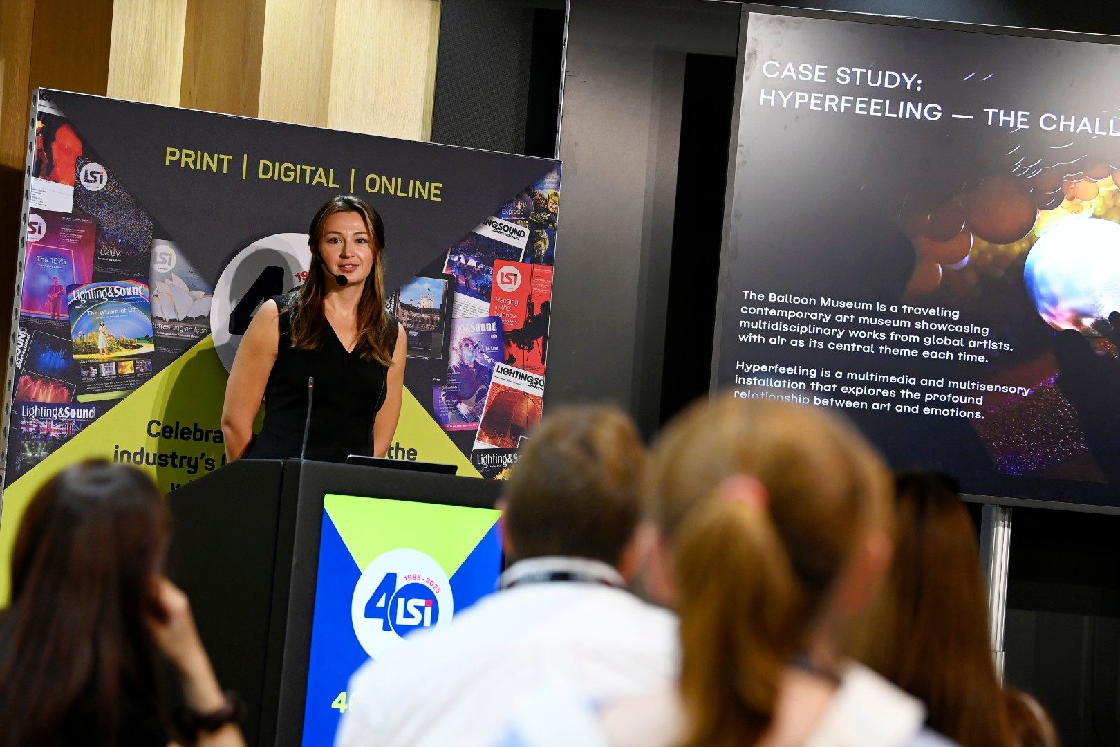 A female speaker with long brown hair presenting a case study titled ‘Hyperfeeling - The Challenge’ during an LSi Theatre session at PLASA Show London 2025, discussing the Balloon Museum’s multisensory art installation.