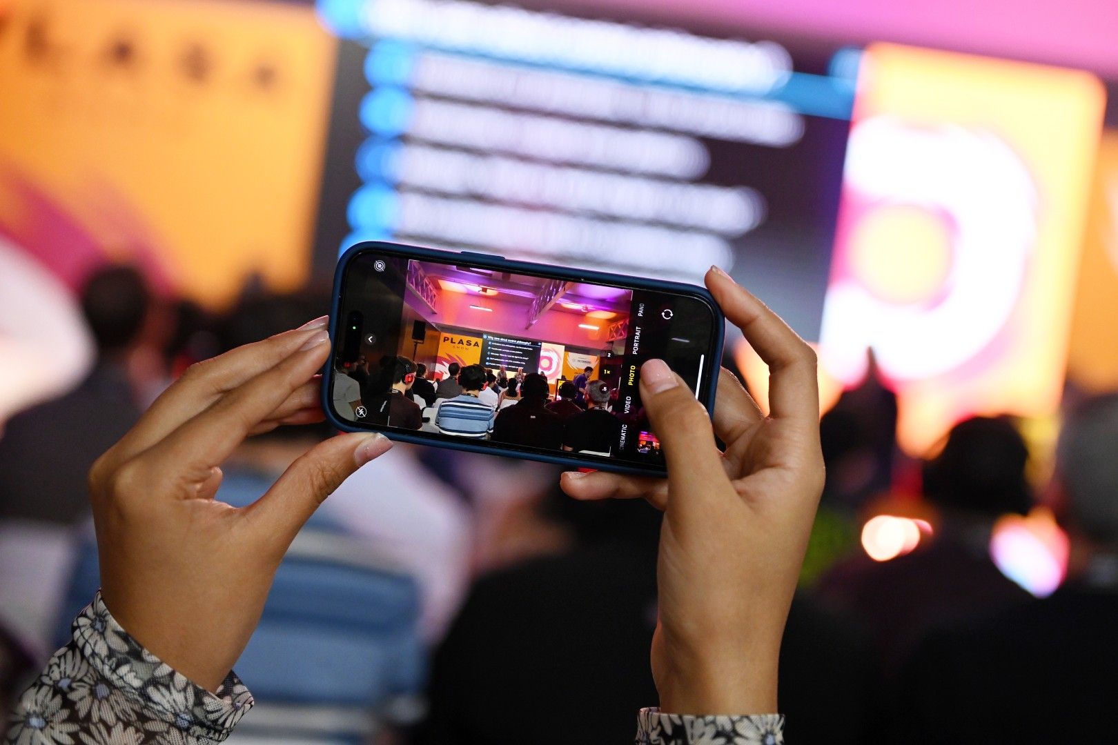 Close-up of a person taking a photo of a seminar audience on a smartphone at PLASA Show London 2025, with colourful stage lighting and presentation screens in the background