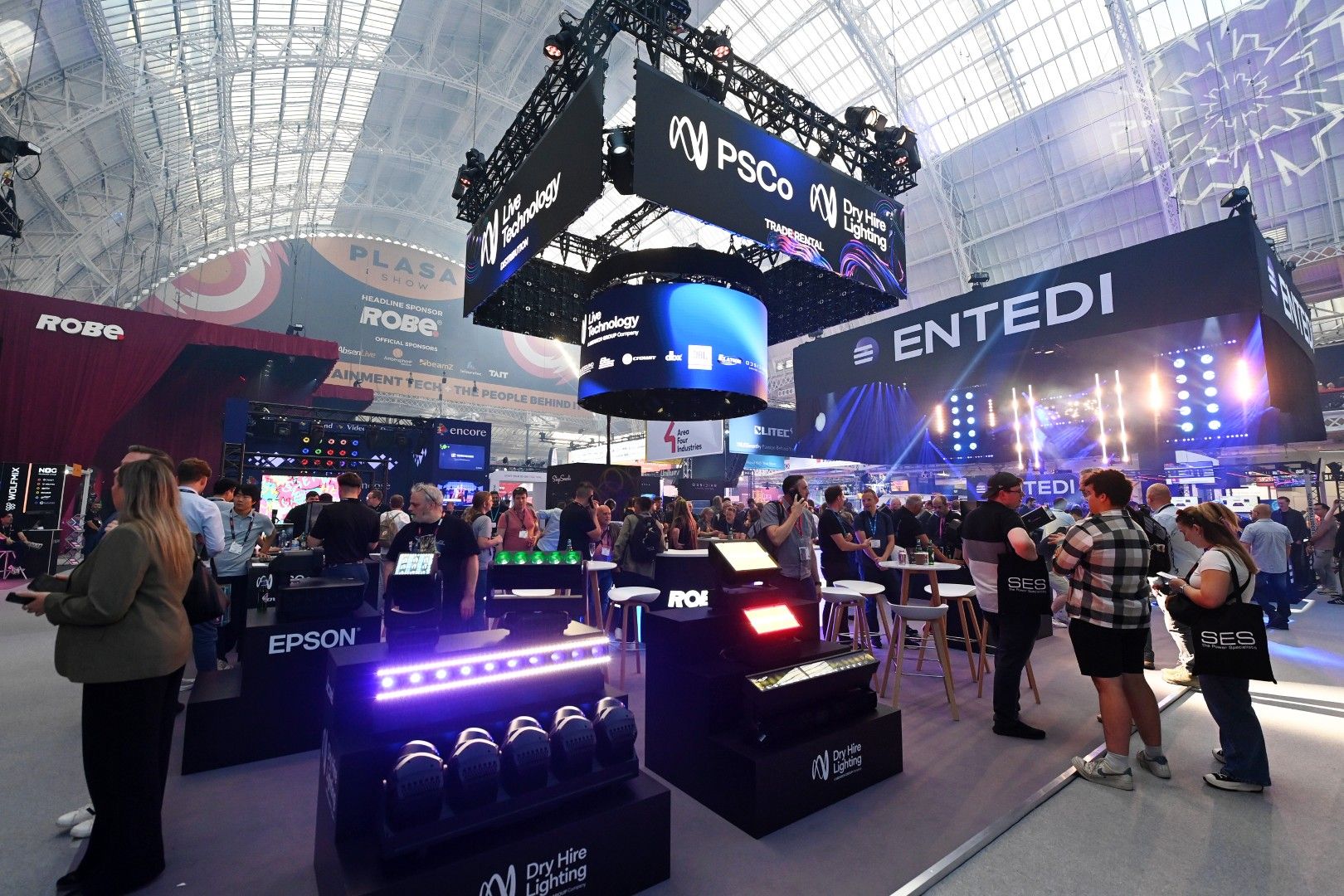Busy exhibition floor at PLASA Show London 2025 inside Olympia, featuring the ENTEDI stand with a lit stage display, the PSCo and Dry Hire Lighting rig above.