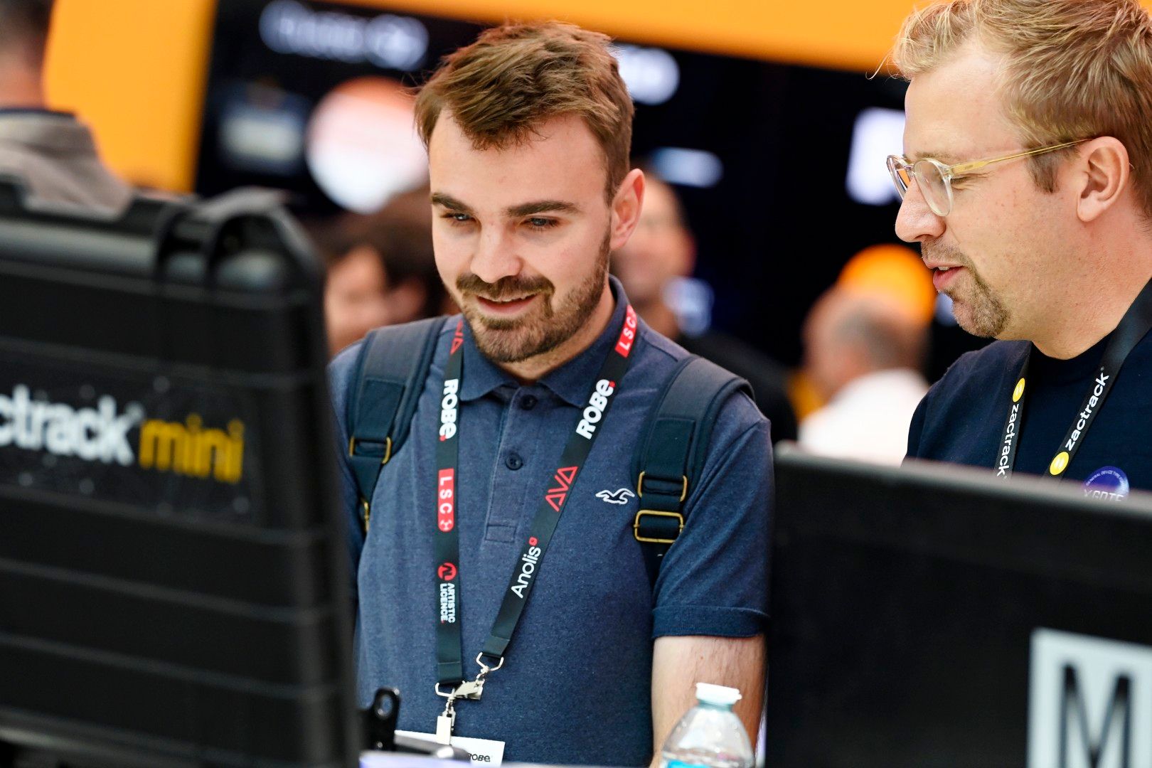 Two men engage in a technical discussion at the zactrack mini stand during PLASA Show London 2025, with one visitor wearing a backpack and event lanyard.