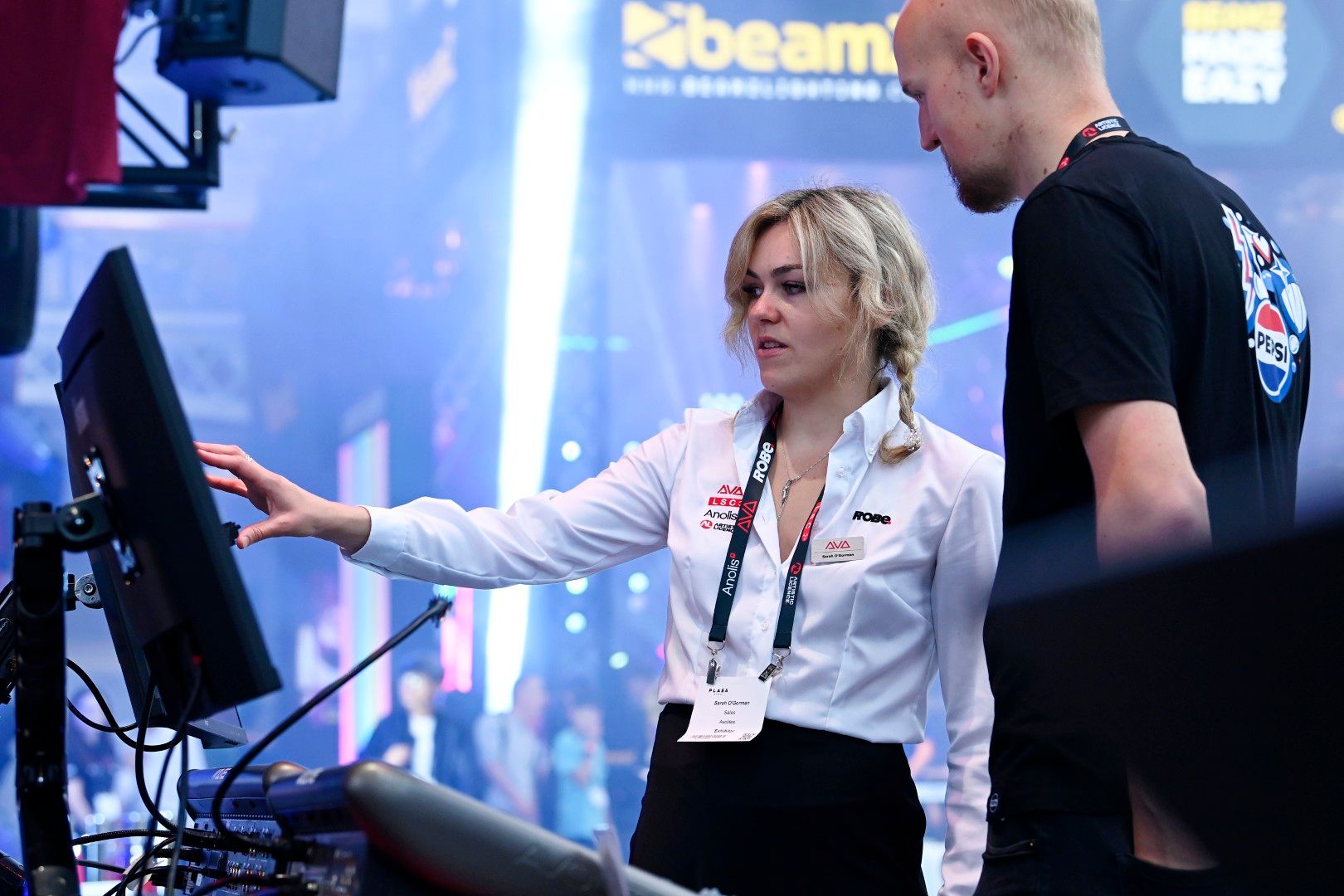 A blonde woman in a white shirt demonstrates a lighting control system to a man at the Robe stand during PLASA Show London 2025.