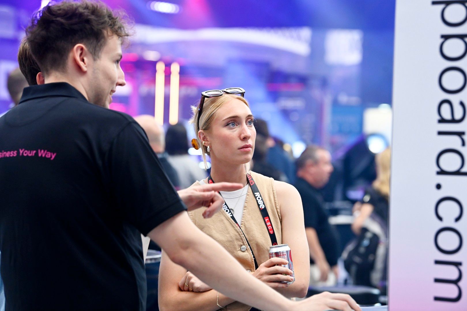 Female visitor with blonde hair listens attentively to an exhibitor’s explanation at PLASA Show London 2025, holding a drink can on the busy show floor.
