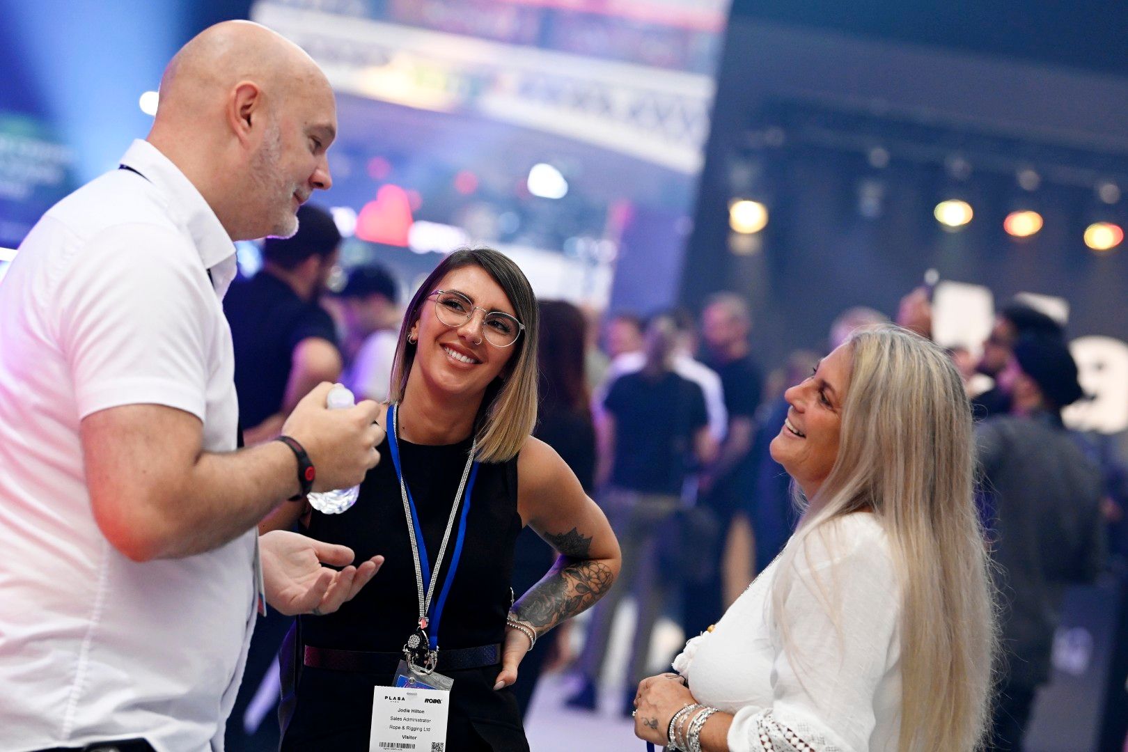 Two women and a man networking and smiling on the show floor at PLASA Show London 2025, with stage lighting visible in the background