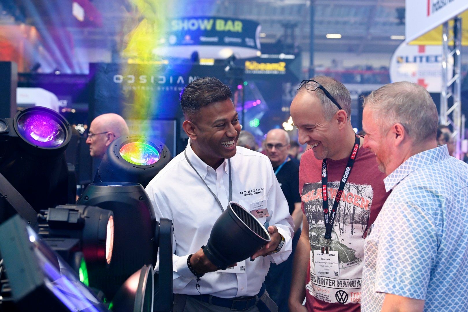 Male exhibitor with short dark hair in a white Obsidian Control Systems shirt demonstrating a professional lighting fixture to two male visitors at PLASA Show London 2025, surrounded by colourful stage lights on the show floor