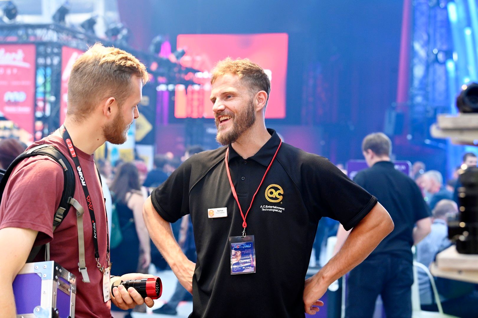 Male exhibitor in a black A.C. Entertainment Technologies shirt smiling and talking with a male visitor holding a lighting connector at PLASA Show London 2025