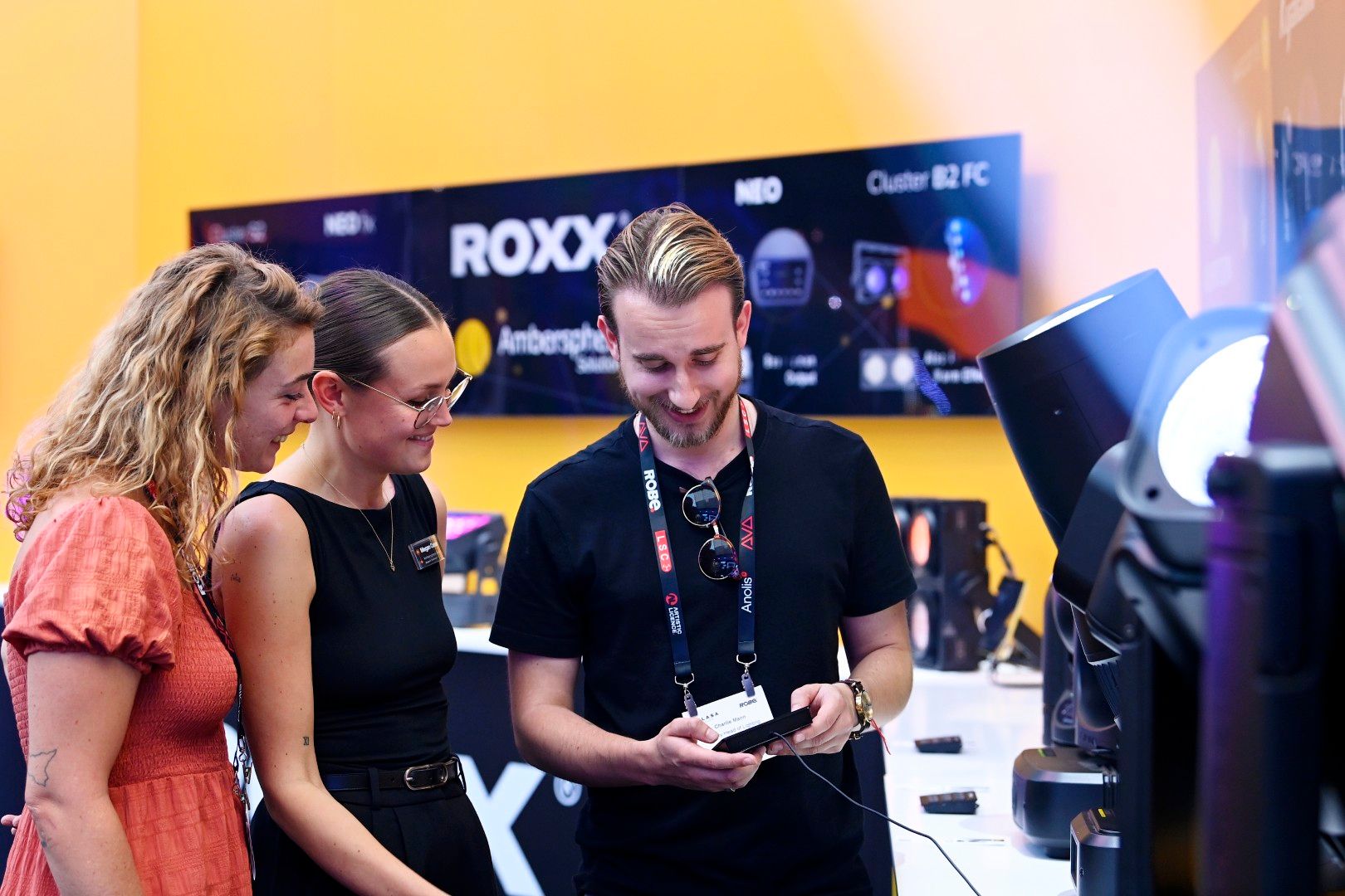Male exhibitor in a black shirt demonstrating lighting control equipment to two female visitors at the Ambersphere stand during PLASA Show London 2025, with illuminated fixtures and product displays in the background