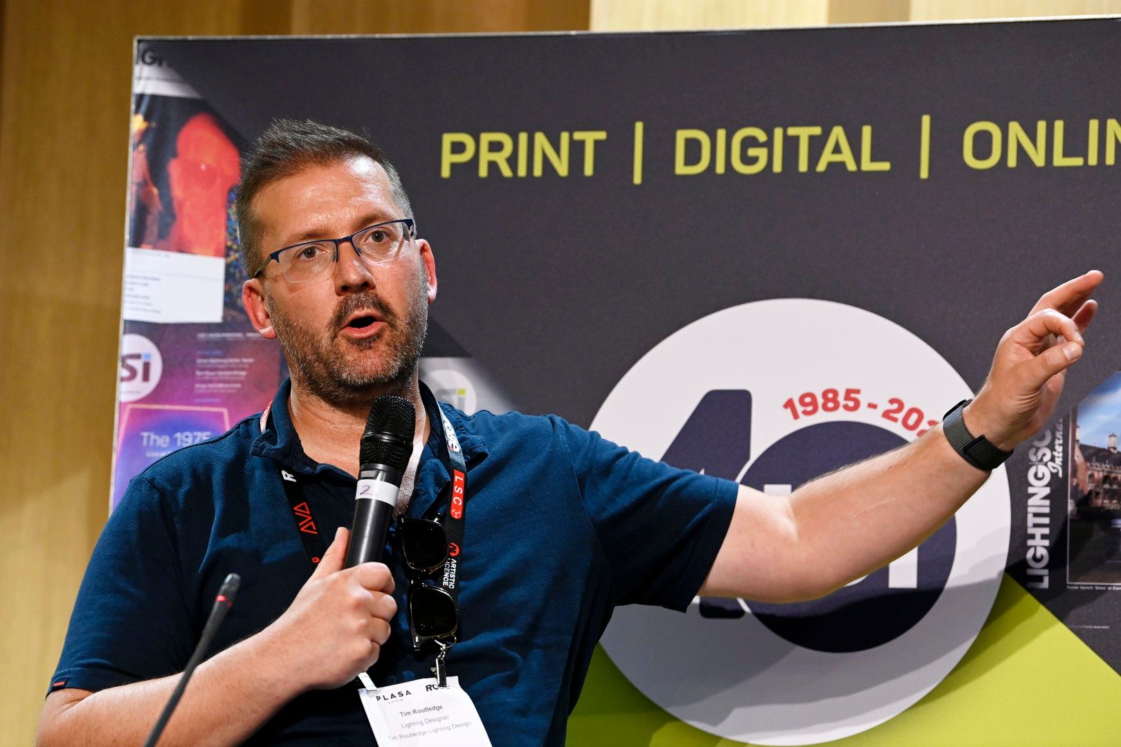 Male lighting designer Tim Routledge, wearing glasses and a navy shirt, speaking into a microphone and gesturing during a seminar at PLASA Show London 2025, with LSi Magazine 40th anniversary branding behind him
