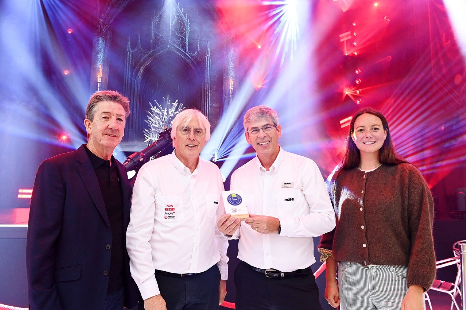 Representatives from Robe Lighting holding a carbon reduction commitment plaque at PLASA Show London 2025, standing in front of a stage with colourful lighting beams and scenic backdrop