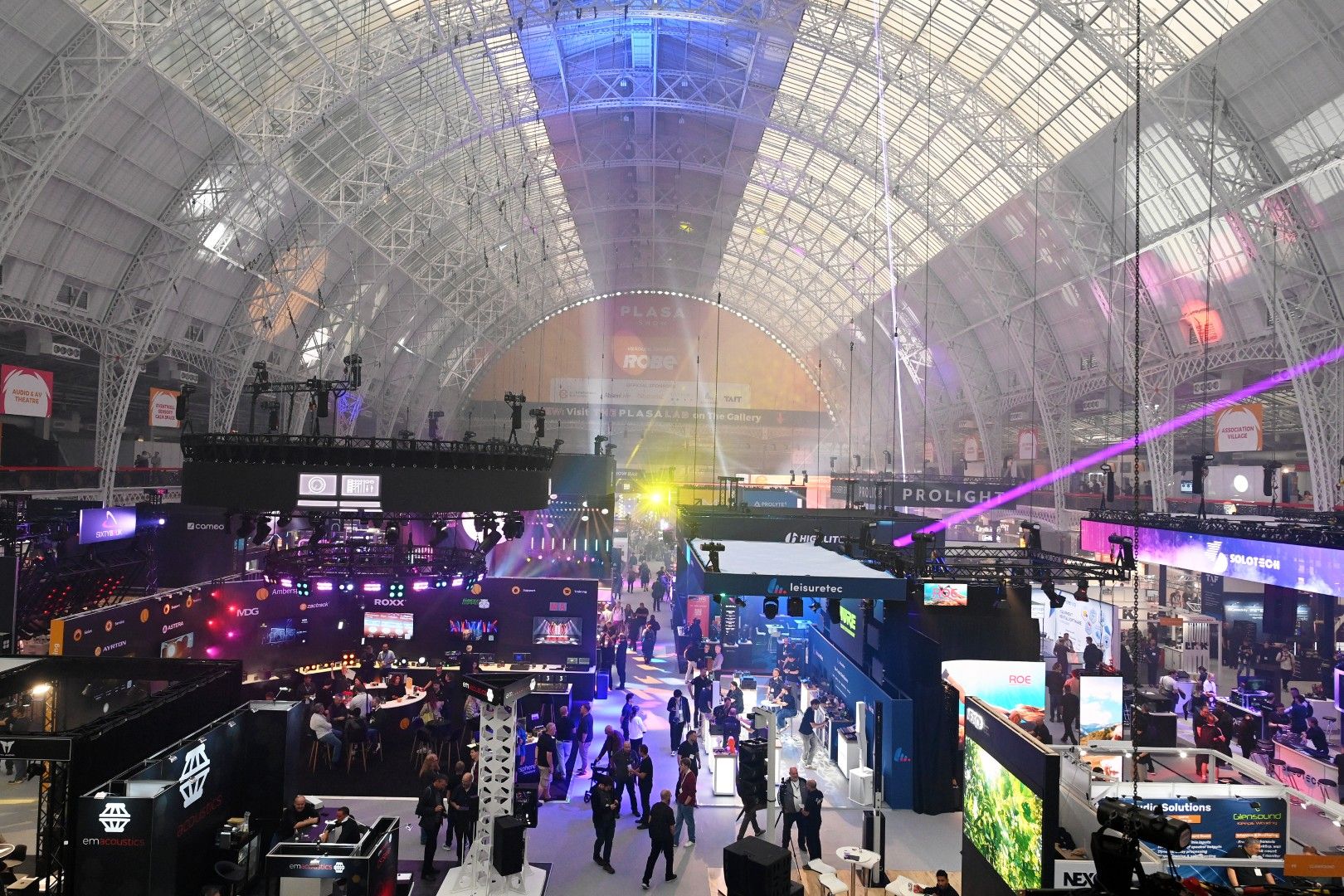 Wide view of the busy PLASA Show 2026 exhibition floor at Olympia London, with visitors exploring entertainment technology stands beneath lighting beams, suspended truss and the venue’s glass roof.