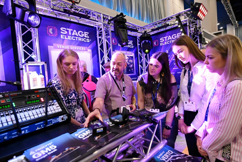 One man and three young women gathered at the Stage Electrics stand, watching a demonstrator explain audio and lighting equipment on a truss-mounted display at PLASA Focus Leeds.