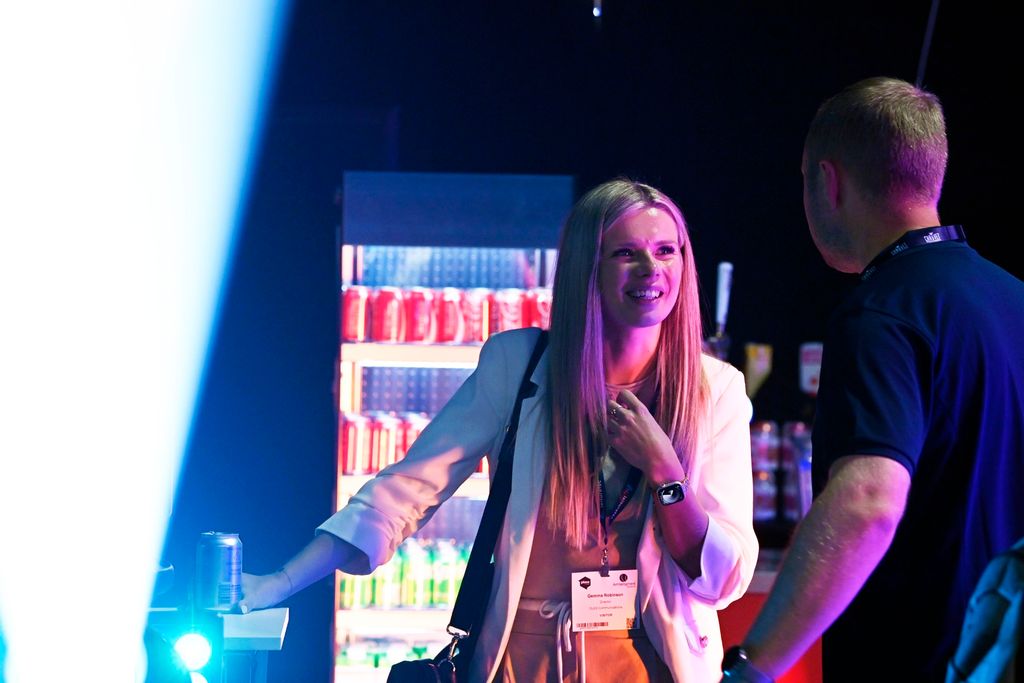 Woman and man chatting at an exhibition bar area, smiling under colourful stage lighting with illuminated drinks fridge in the background.