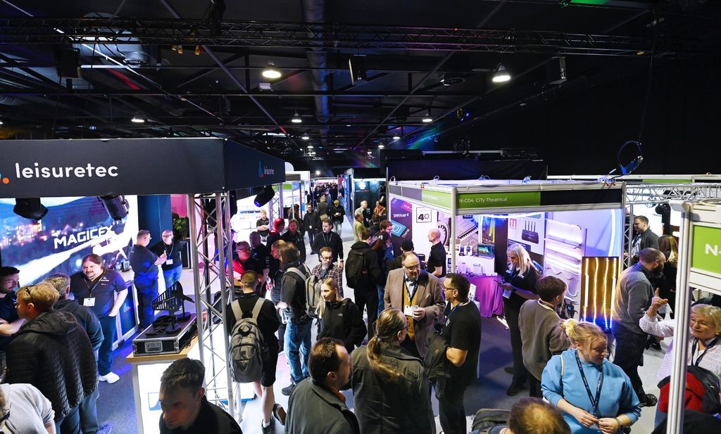 Visitors explore exhibitor stands and network on a busy exhibition floor at PLASA Focus Leeds, surrounded by lighting displays and truss structures